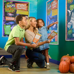 A woman with two children kneels in front of sea creature posters. The young girl points while holding a map. Several pumpkins are on the floor.