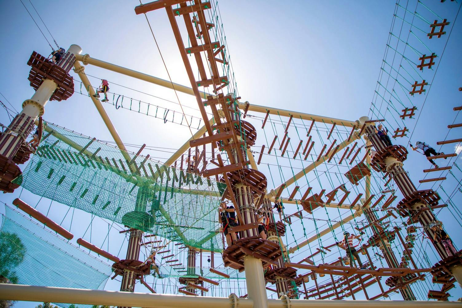 맑고 푸른 하늘을 배경으로 다양한 나무와 밧줄 다리가 있는 높은 밧줄 코스를 항해하는 사람들이 많습니다.