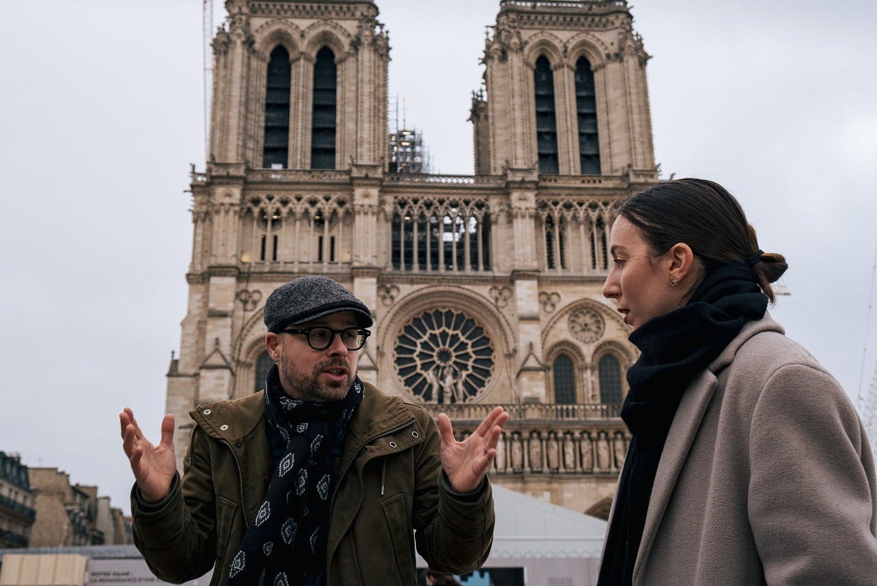 Two people talking in front of a gothic cathedral with ornate architecture and large rose window.
