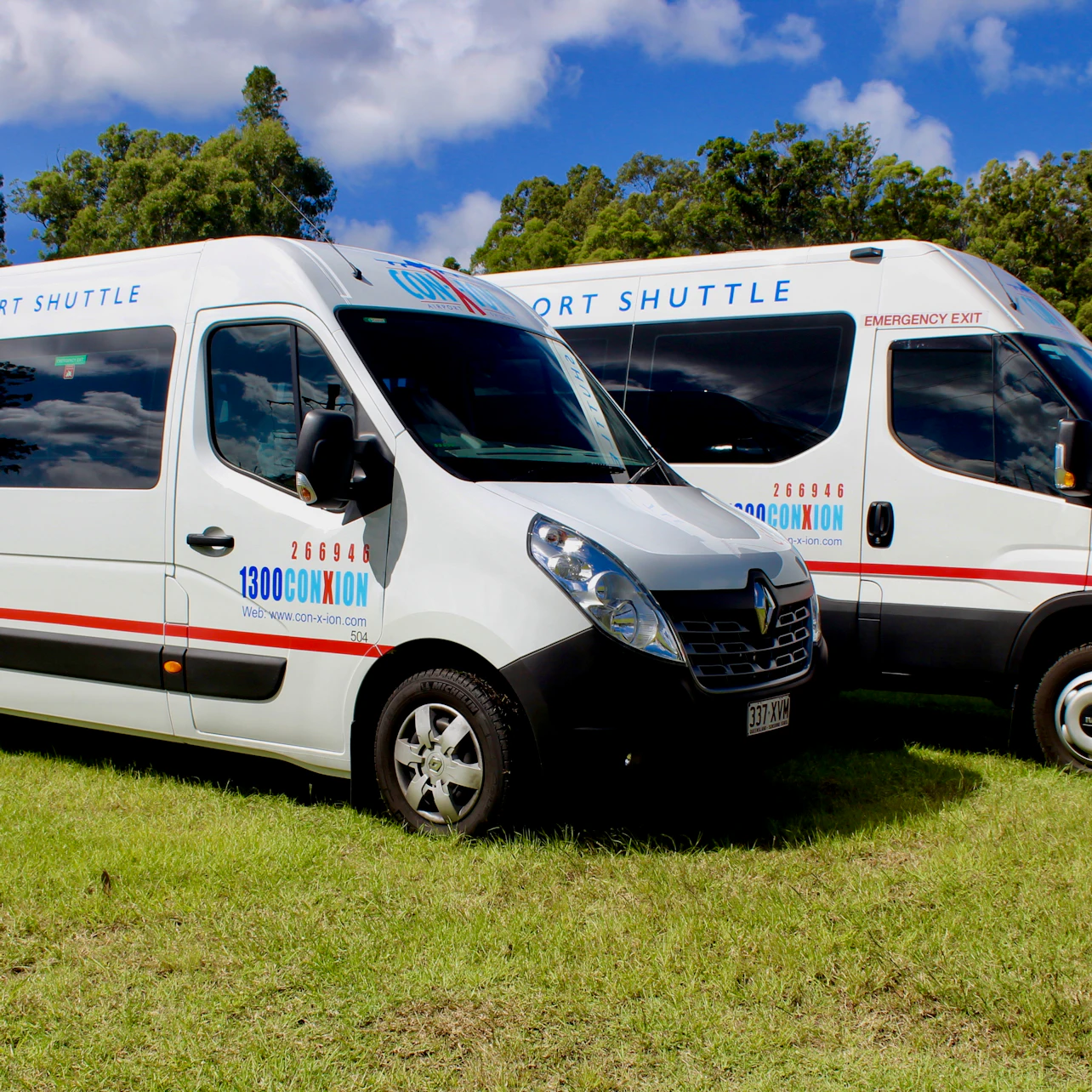 Two white transport shuttles with blue and red lettering parked on grass under a blue sky with trees in the background.