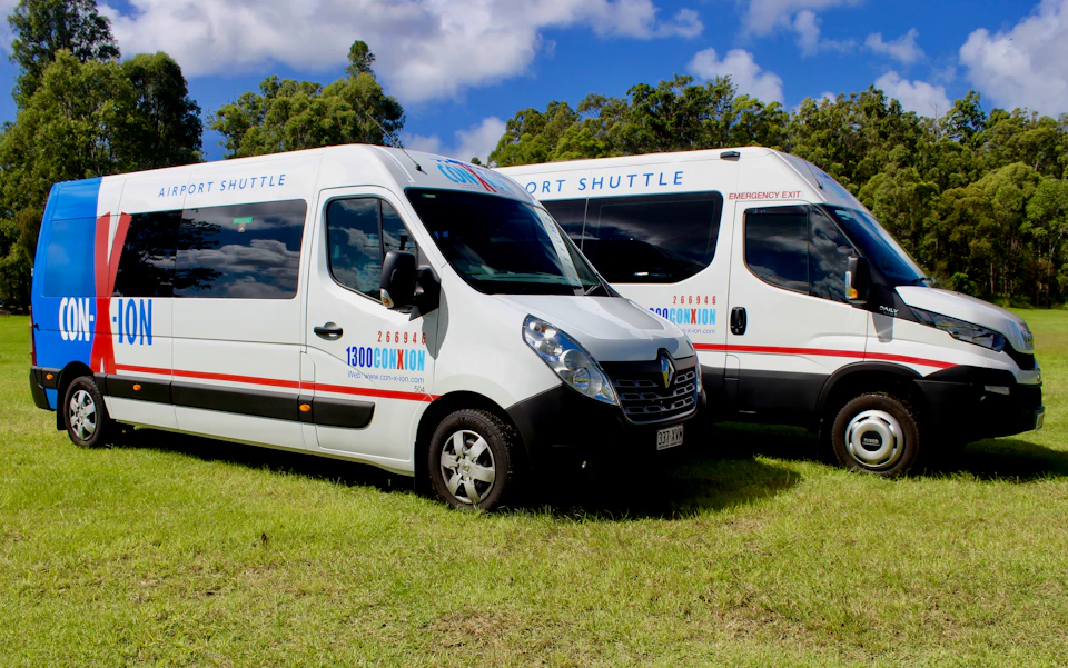 Two white shuttle vans with "100% ConXion" branding, parked on grass under a partly cloudy sky.