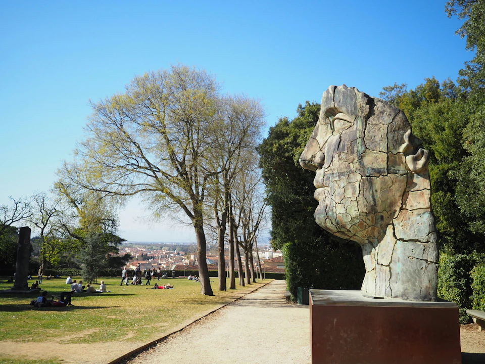 A large cracked face sculpture beside a path with trees and people sitting on a lawn; city in the distant background.