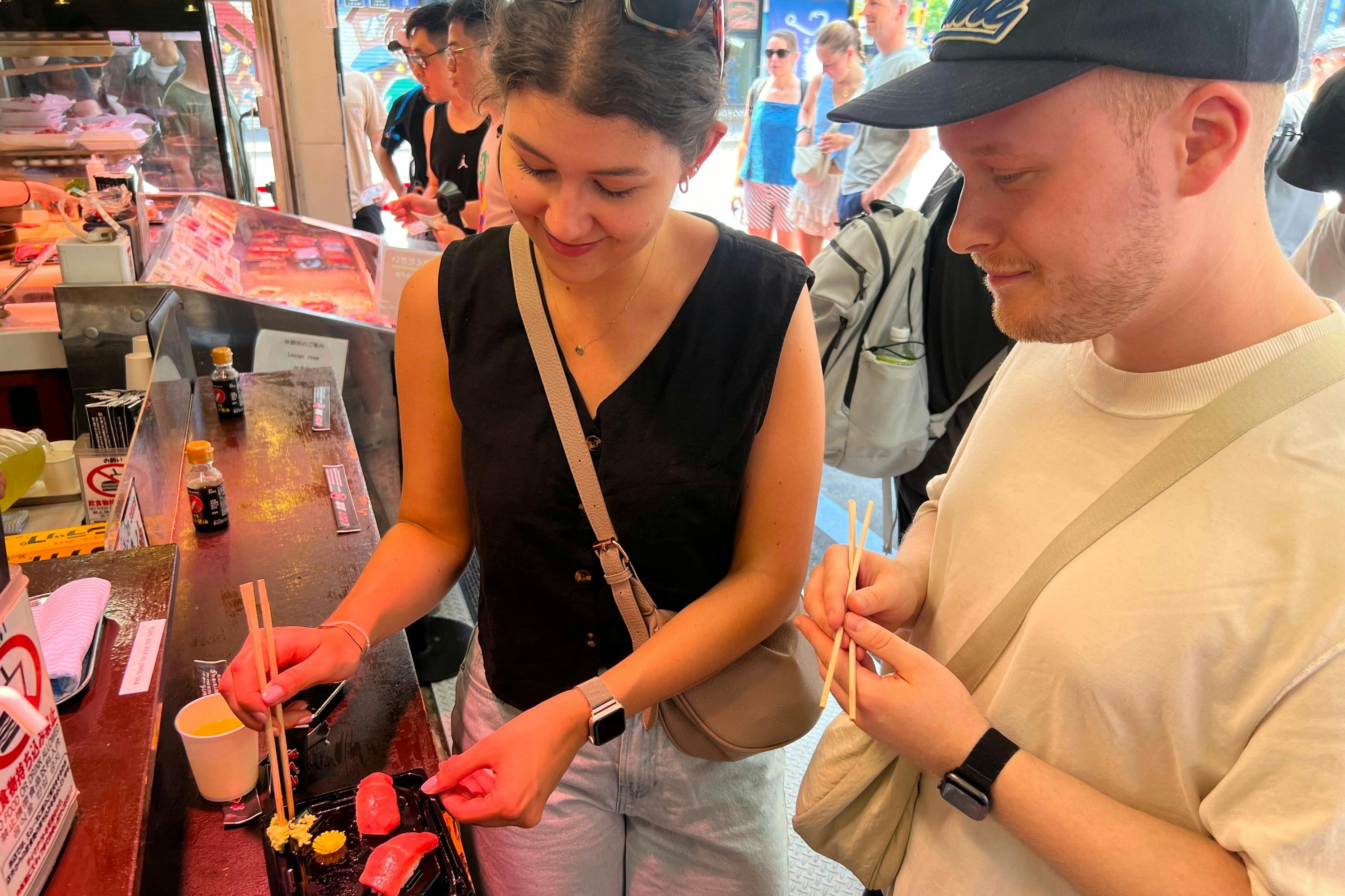 Two people standing closely together, one holding chopsticks, in a casual setting with others visible in the background.