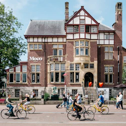 Människor cyklar och går framför Moco Museum, en stor tegelbyggnad med detaljerad arkitektur och skyltar för Banksy och Dalí.