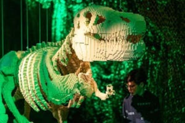 Ett stort dinosaurieskelett av LEGO-klossar som visas under grön belysning, med en person som står i närheten och observerar det.