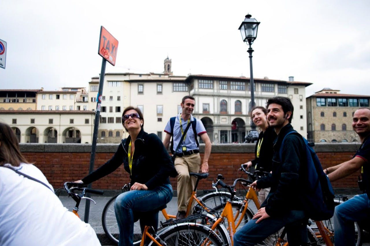 People on bicycles smiling in front of buildings, with a sign reading "Taxi" on a pole nearby.