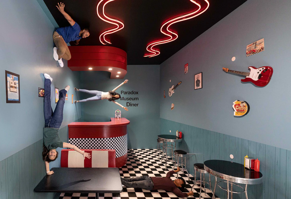 People appear to be standing sideways and upside down in a diner with a checkered floor, red counter, and teal walls adorned with guitars.
