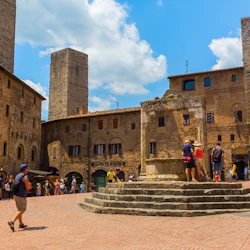 Människor samlas kring en gammal stenbrunn på ett historiskt torg, omgivet av gamla tegelbyggnader och höga stentorn.