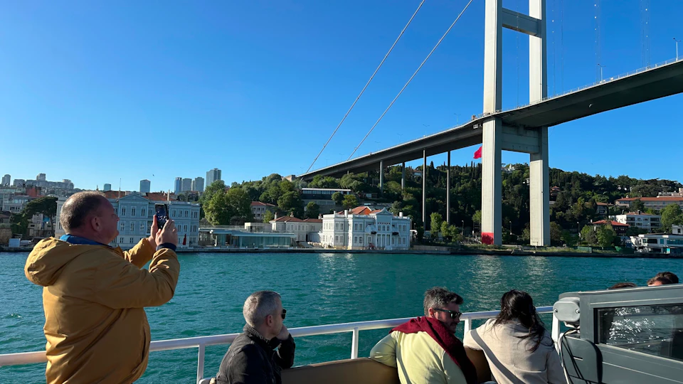 People on a boat, with a view of a large suspension bridge, waterfront buildings, and green hills under a clear blue sky.