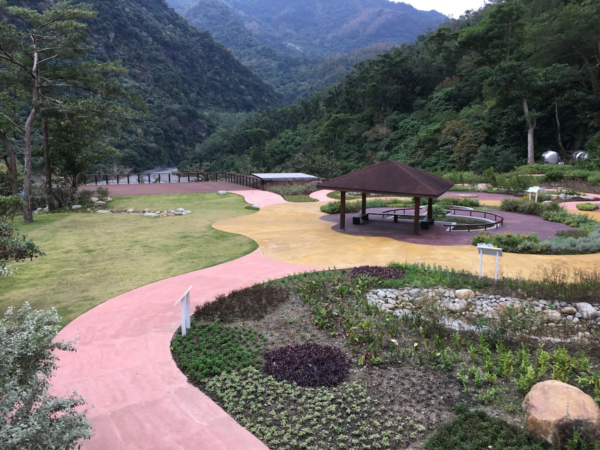 A landscaped park with pink walkways, a gazebo, plants, and hills in the background under an overcast sky.