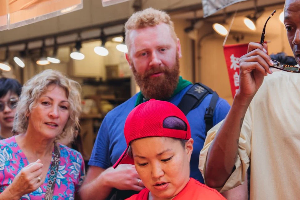 A group of people gather around a food stall. A person in a red shirt appears to be preparing or handling items.