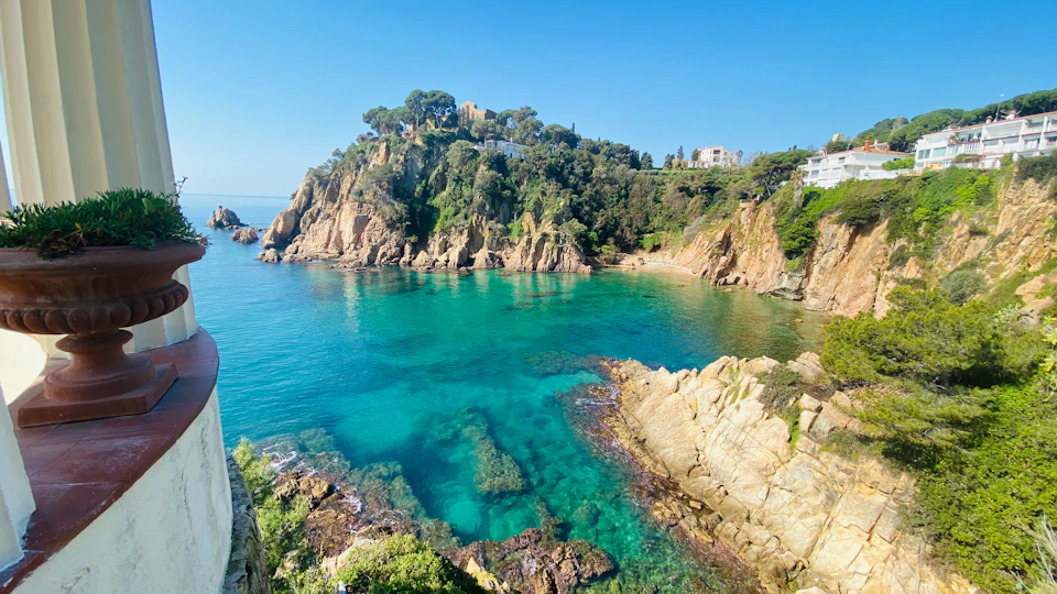 A rocky coastline with clear blue water, rugged cliffs, and buildings nestled among trees under a clear blue sky.