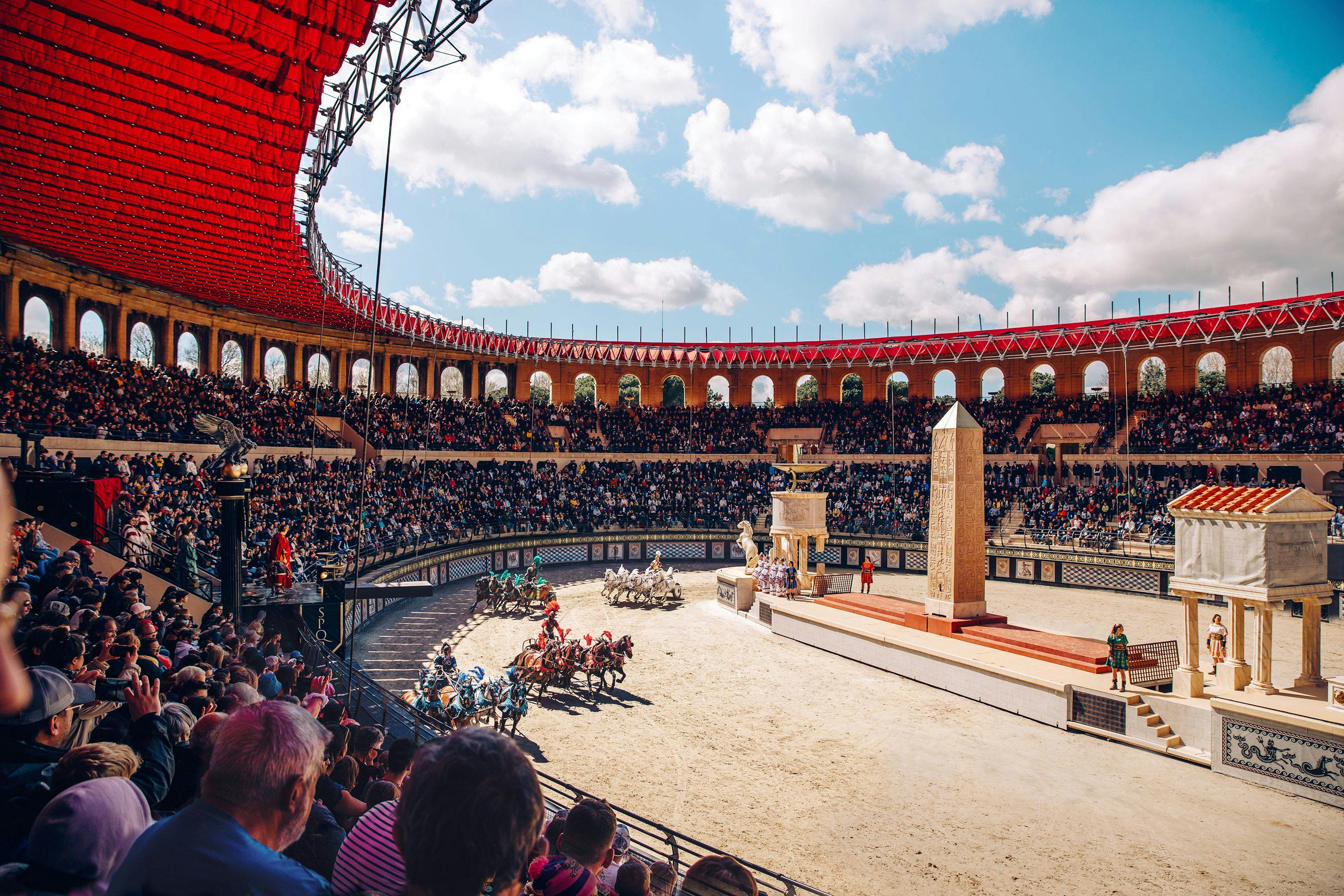 Ein von Pferden gezogenes Wagenrennen in einer großen Arena mit gestaffelten Sitzplätzen, die mit Zuschauern unter einem teilweise überdachten Himmel gefüllt ist.