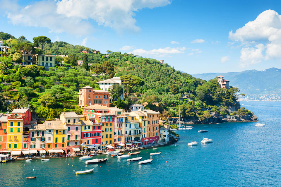 Colorful buildings line the waterfront of a coastal town with boats anchored in the blue water and green hills in the background.