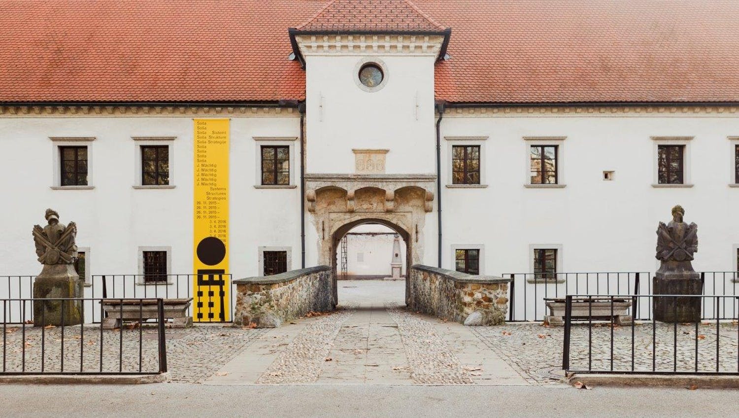 Historická budova s červenou kachlovou střechou, klenutým vchodem a žlutým informačním panelem na levé straně.