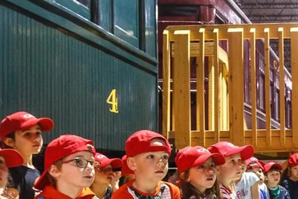 A group of children wearing red caps sits on a bench inside a train museum, with vintage train cars on either side.