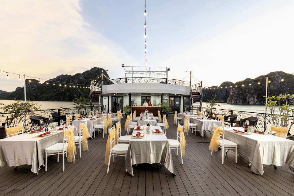 An outdoor dining area on a boat, decorated with tables, chairs, and string lights, set against a backdrop of water and cliffs.
