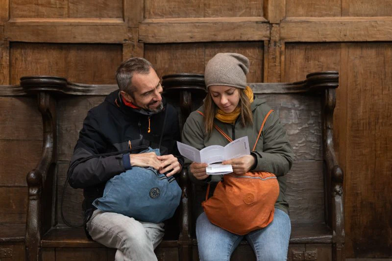 Two people sit on a wooden bench. One reads a booklet while the other looks on. Both carry backpacks.