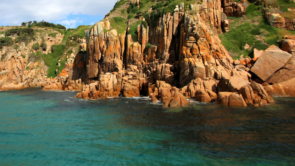 Rugged, orange-brown cliffs with patches of green vegetation rise above clear turquoise water.