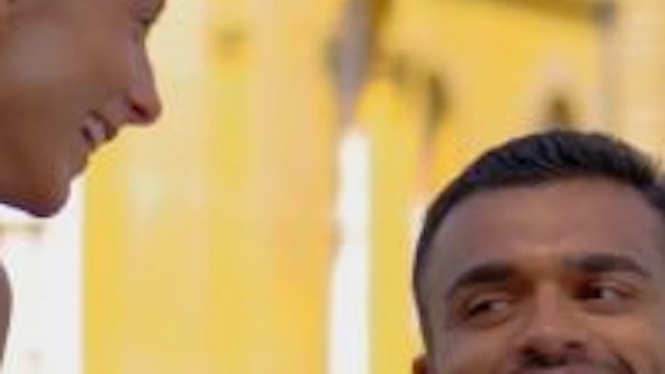 A couple sits on a red picnic blanket on a green lawn, smiling at each other in front of a historic yellow building with towers.