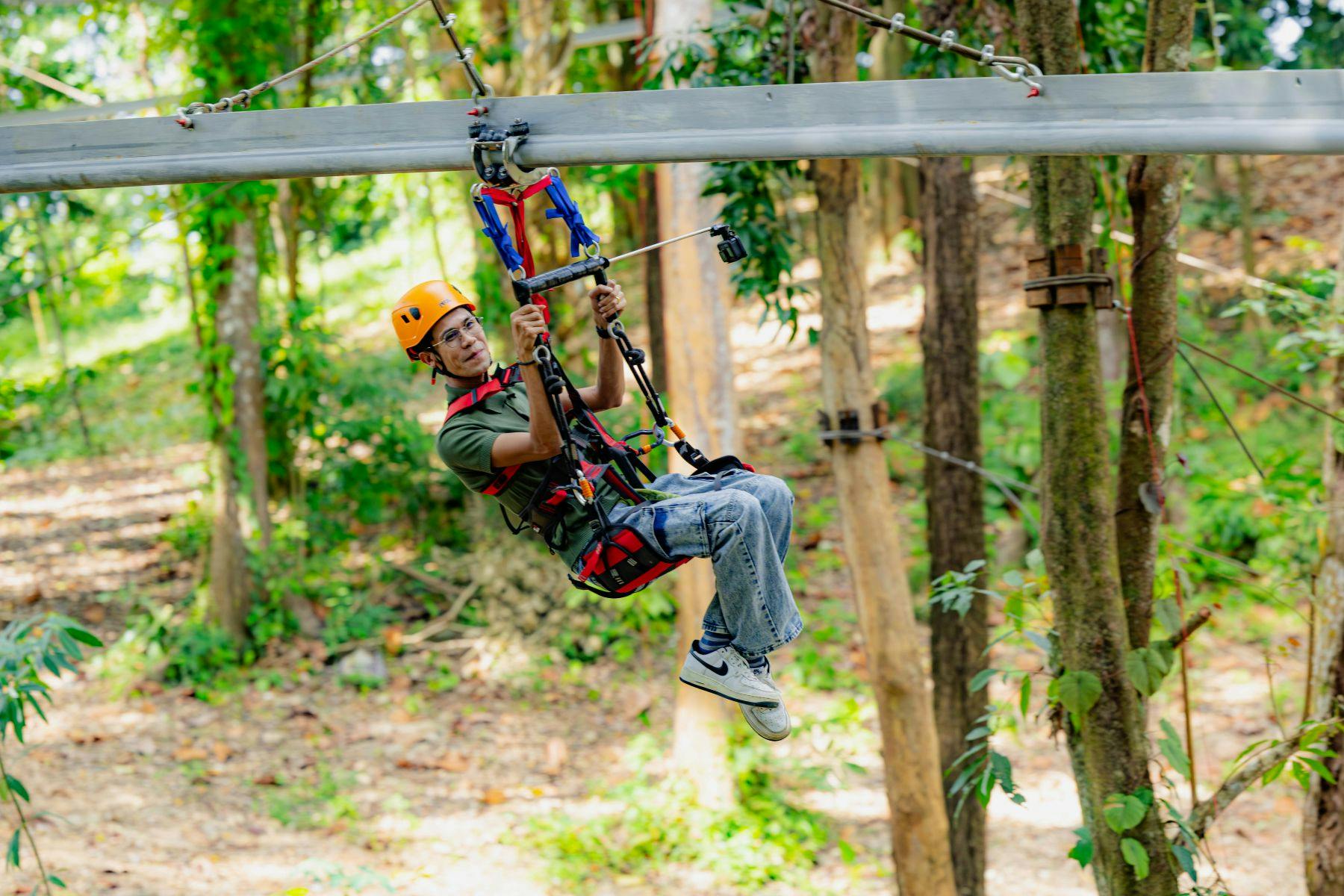 A person wearing safety gear and an orange helmet gliding on a zipline through a forested area.