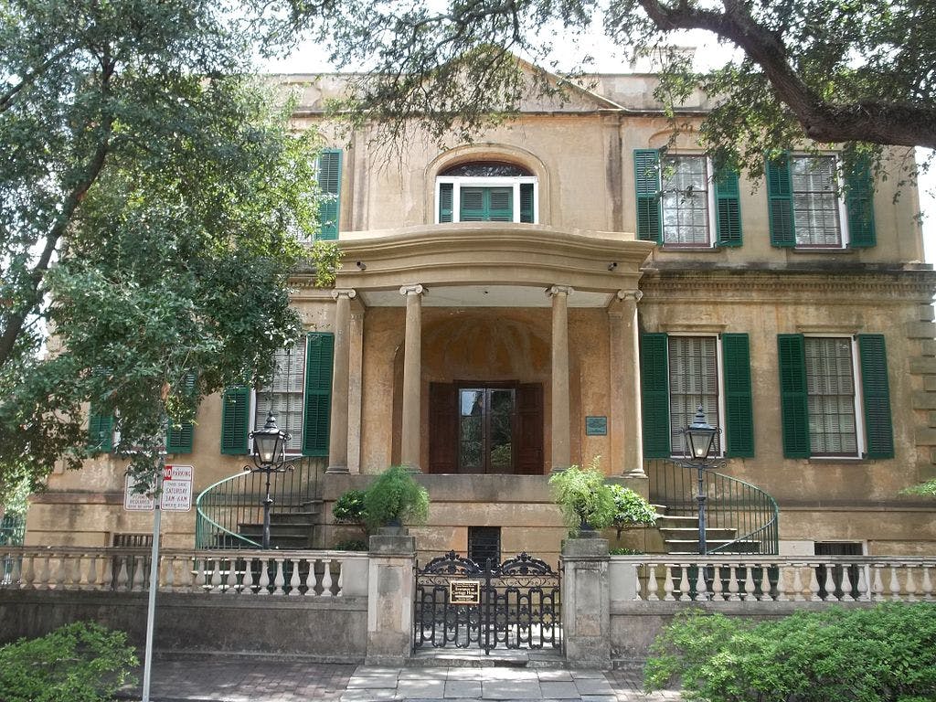 A historic building with green shutters, arched windows, and a grand entrance flanked by columns, surrounded by trees and greenery.