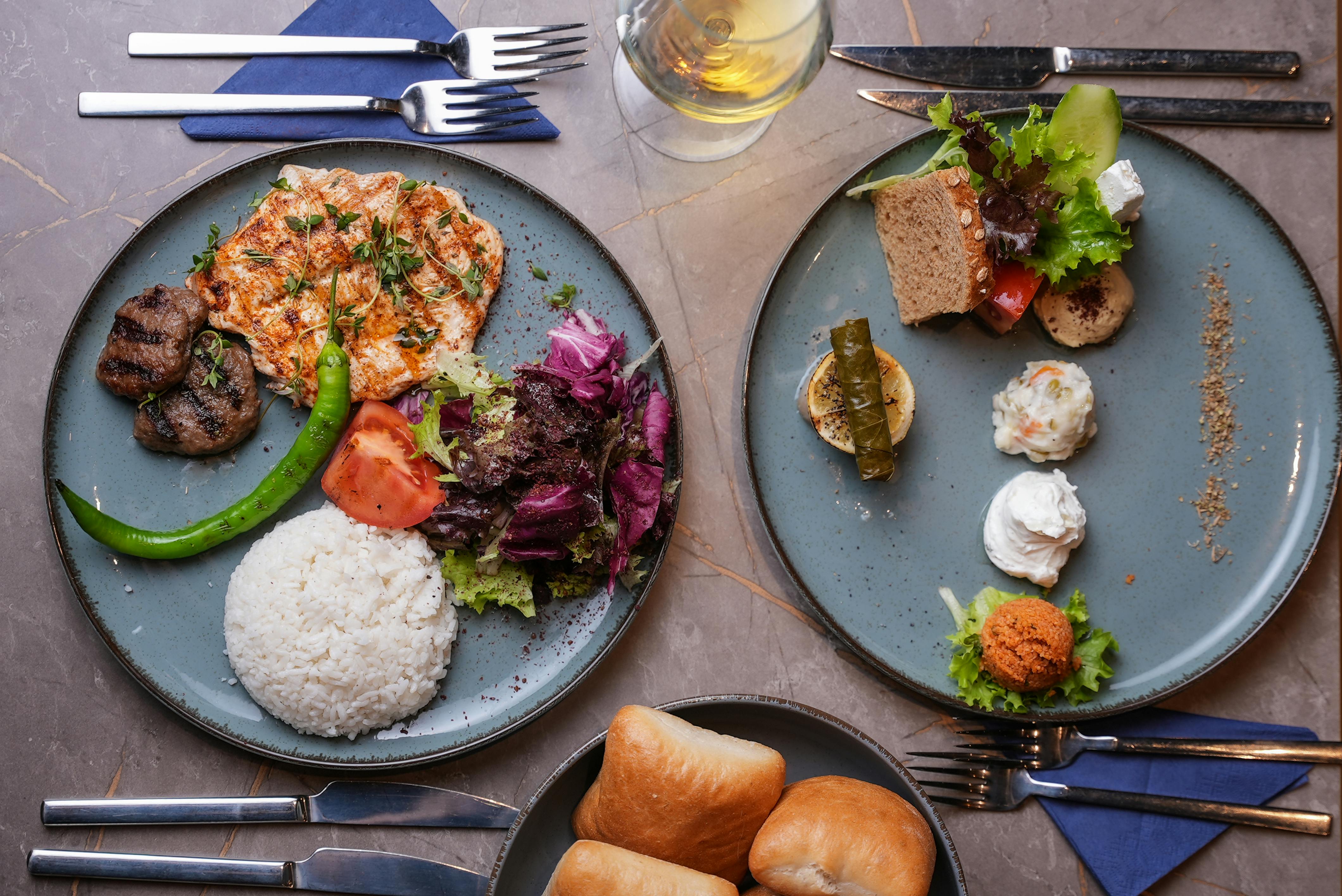 ご飯、鶏肉のグリル、サラダ、パン、前菜を添えた2皿の料理と、テーブルの上にすっきりと並べられたドリンクとカトラリー。