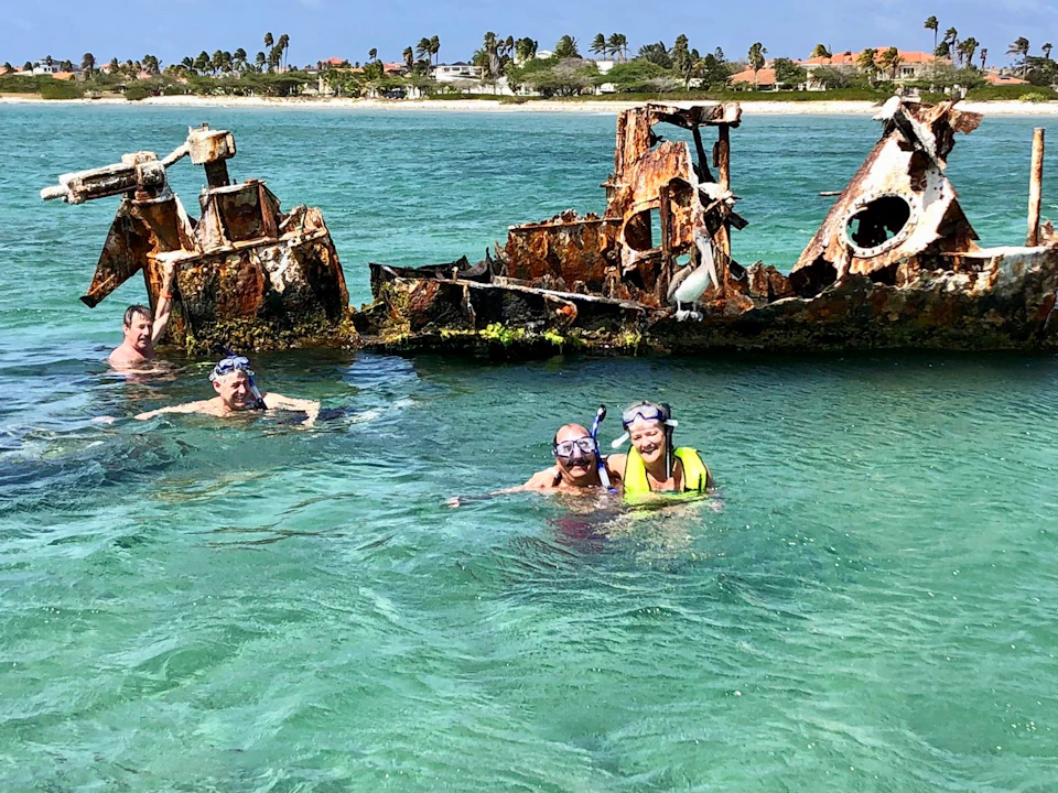 Four people snorkel in clear, turquoise water near a rusted shipwreck with a distant shoreline in the background.