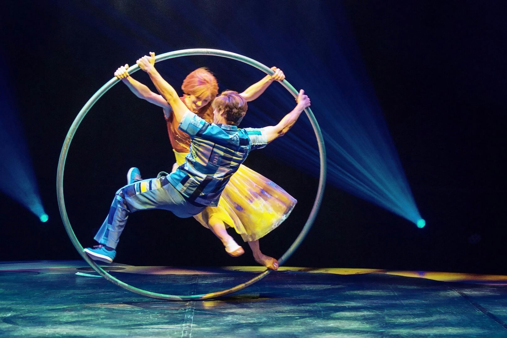 Two performers in colorful outfits balance and spin inside a large metal hoop on a stage with dramatic lighting.