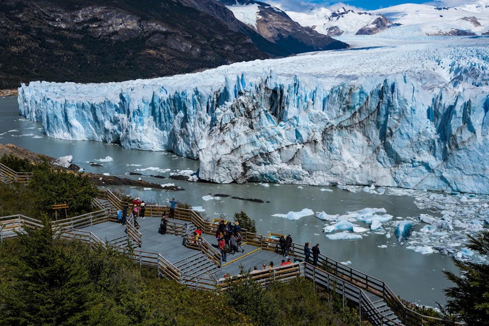 Visitors on wooden walkways observing a massive glacier by a river with mountains in the background.