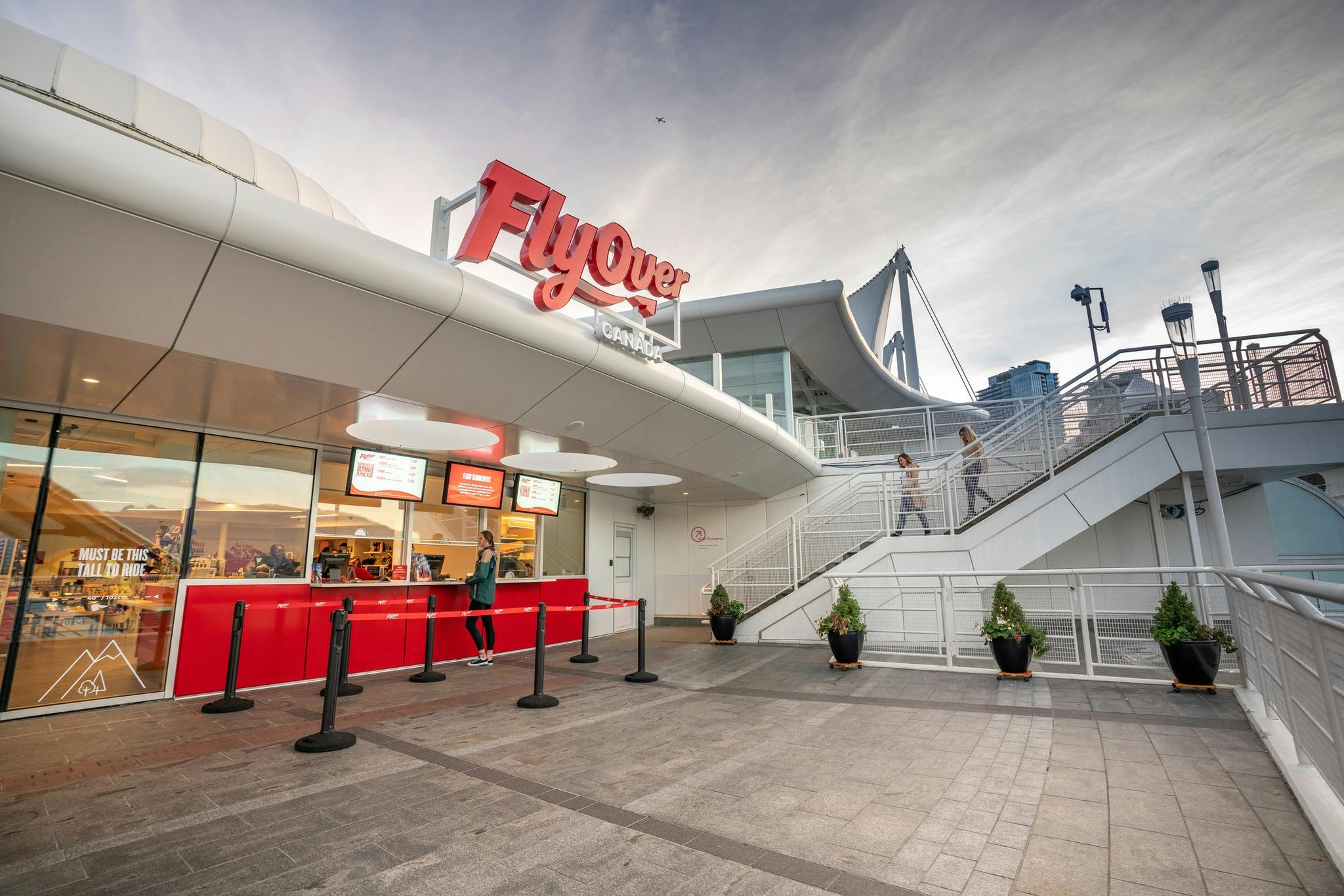 人们站在「飞越加拿大」（ FlyOver Canada ）的入口处，这是一栋现代化的建筑，屋顶弧形，楼梯通向上方。