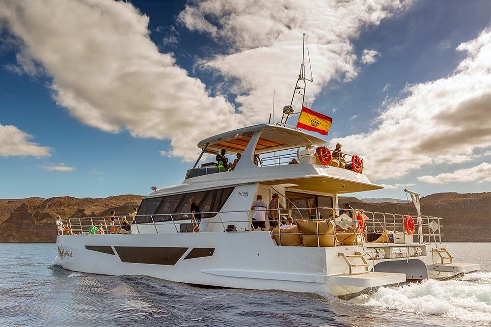 Катамаран с испанским флагом плывет по спокойным водам, пассажиры на палубе наслаждаются пейзажами, скалами на заднем плане под частично облачным небом.