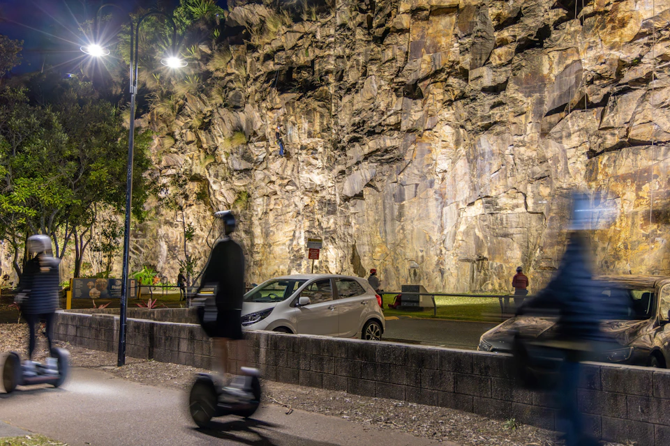 A rock climbing wall at night illuminated by lights, with a person climbing, a car parked nearby, and a person on a scooter in the foreground.