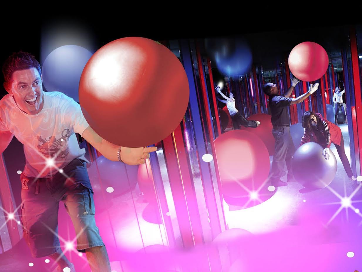 Lächelnde Person, die einen großen, roten aufblasbaren Ball in einem bunten, beleuchteten Raum mit Spiegeln und Reflexionen hält.