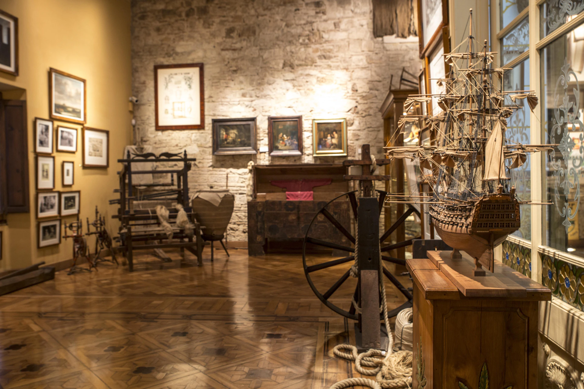 Museum room with wooden floor, ship models, vintage furniture, maritime artifacts, and framed paintings on a stone wall.