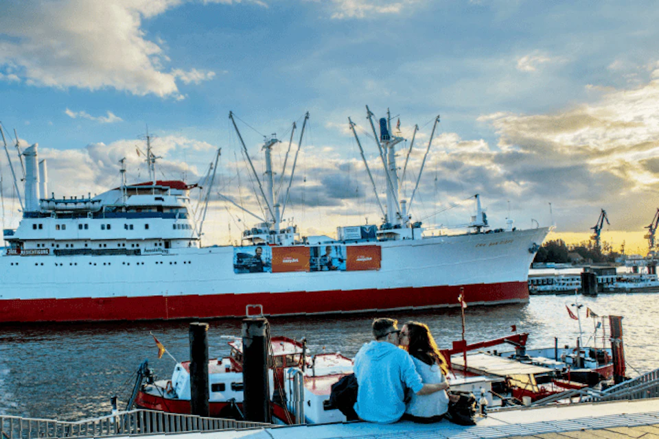 A couple sits on a dock, embracing and facing a large white and red ship under a partly cloudy sky.