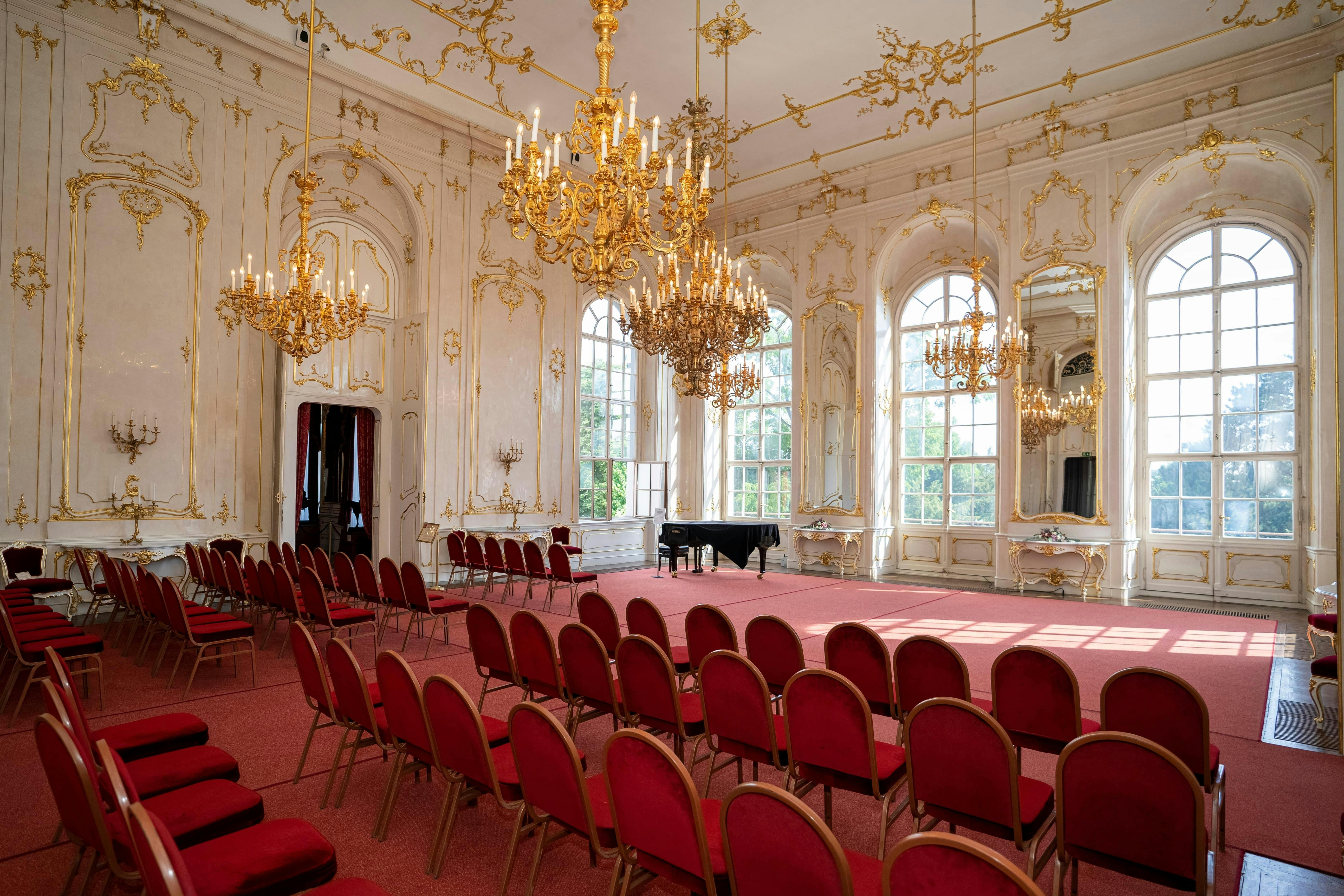 빨간색 의자가 줄지어 있고, 검은색 그랜드 피아노, 커다란 아치형 창문, 높은 천장에 매달린 샹들리에가 있는 화려한 객실입니다.
