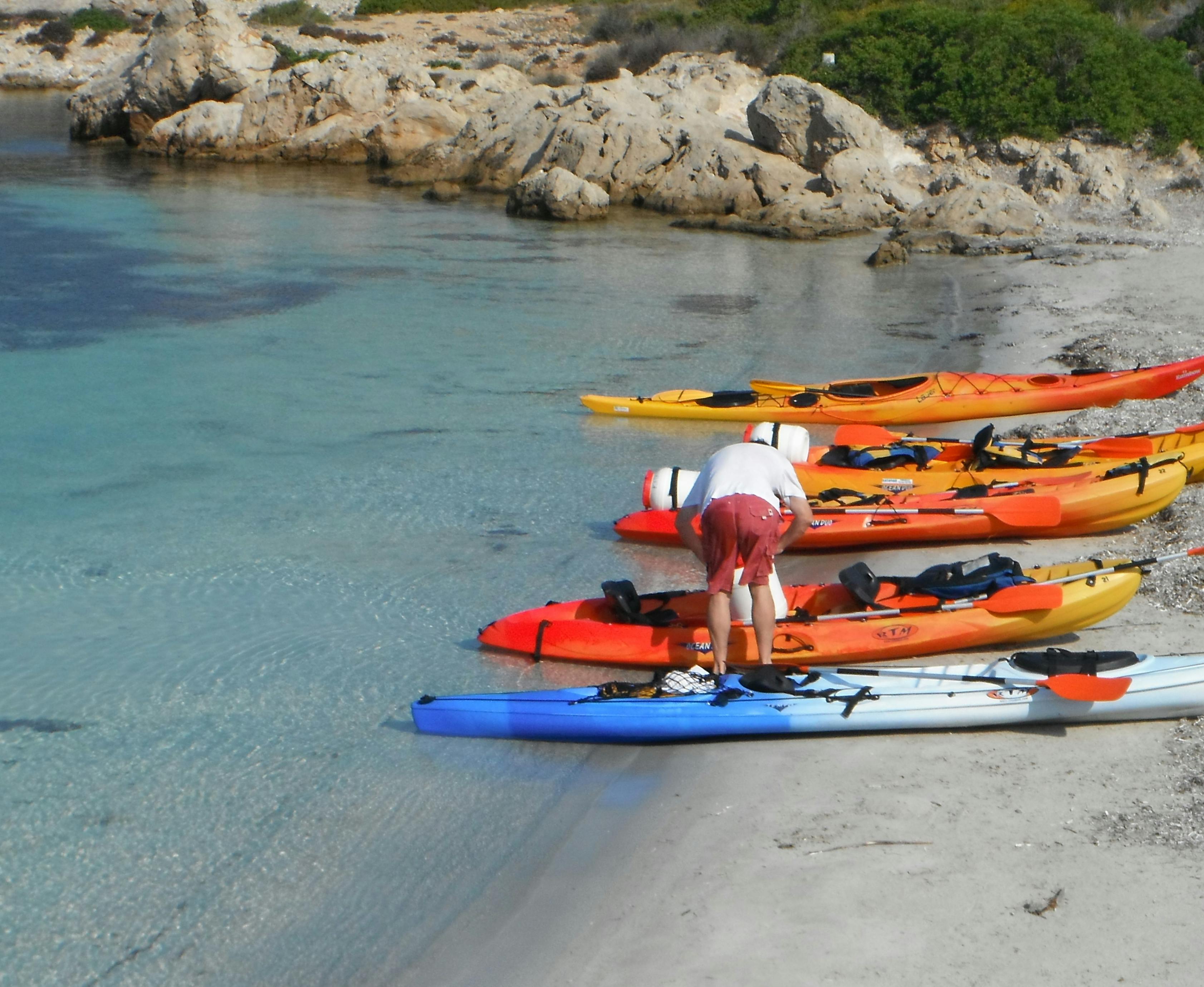 Una persona està parada en una platja de sorra, assegurant objectes en un dels diversos caiacs de colors alineats prop de l'aigua. El terreny rocós és visible al fons.