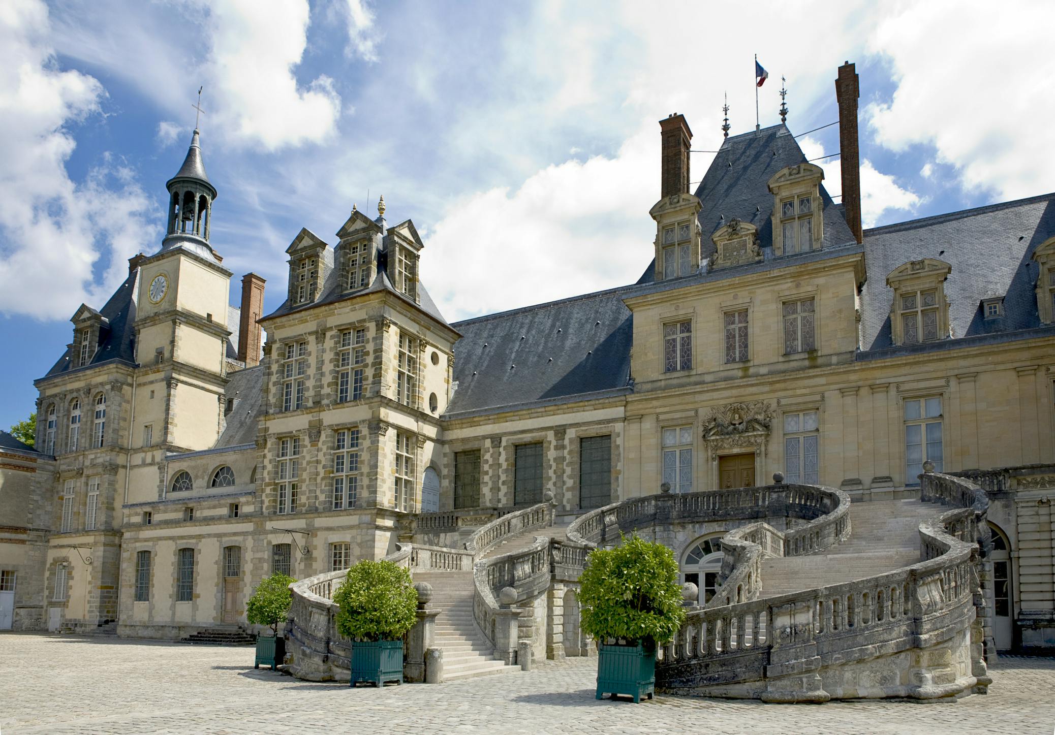 Historic chateau with ornate architecture, tall windows, a grand staircase, and manicured potted plants against a blue sky.