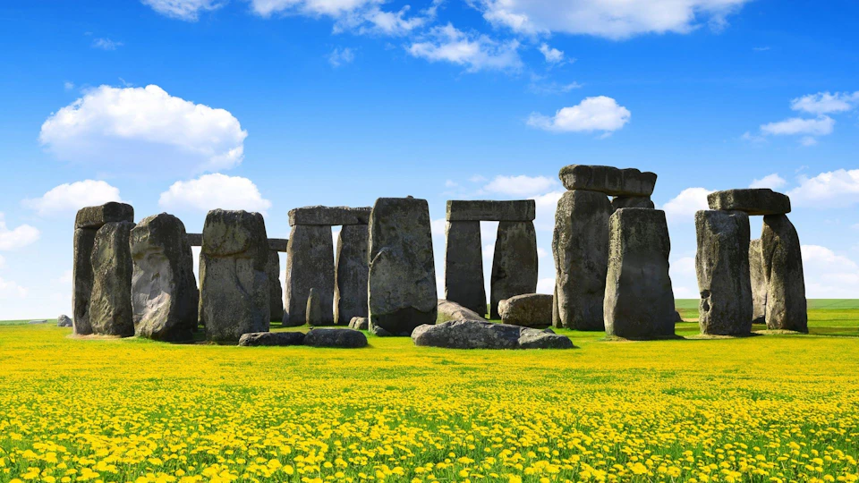 Stonehenge stone circle is surrounded by a field of yellow flowers under a partly cloudy blue sky.