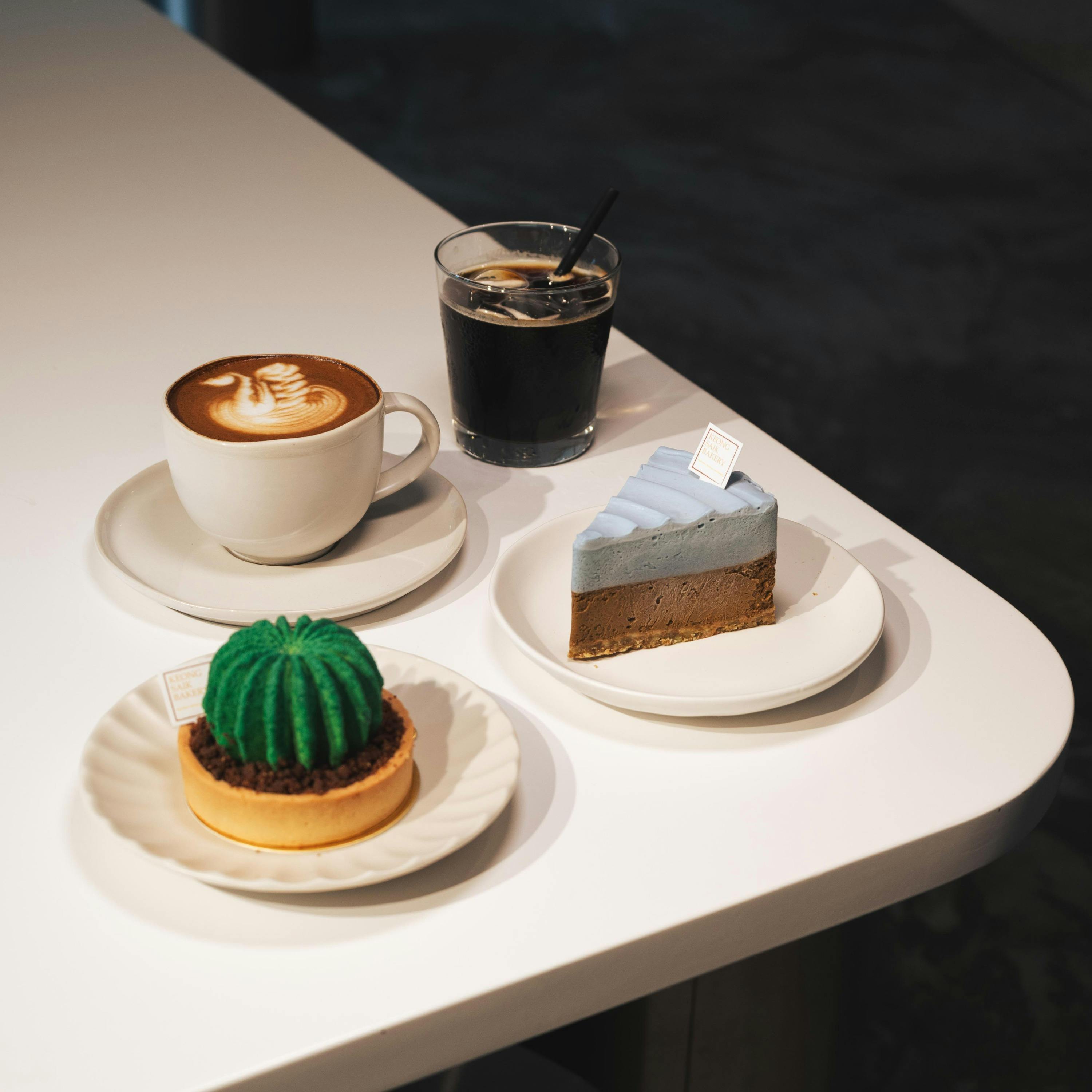 A table with a cup of coffee, a glass of iced coffee, a green dessert, and a slice of blue cake on white plates.