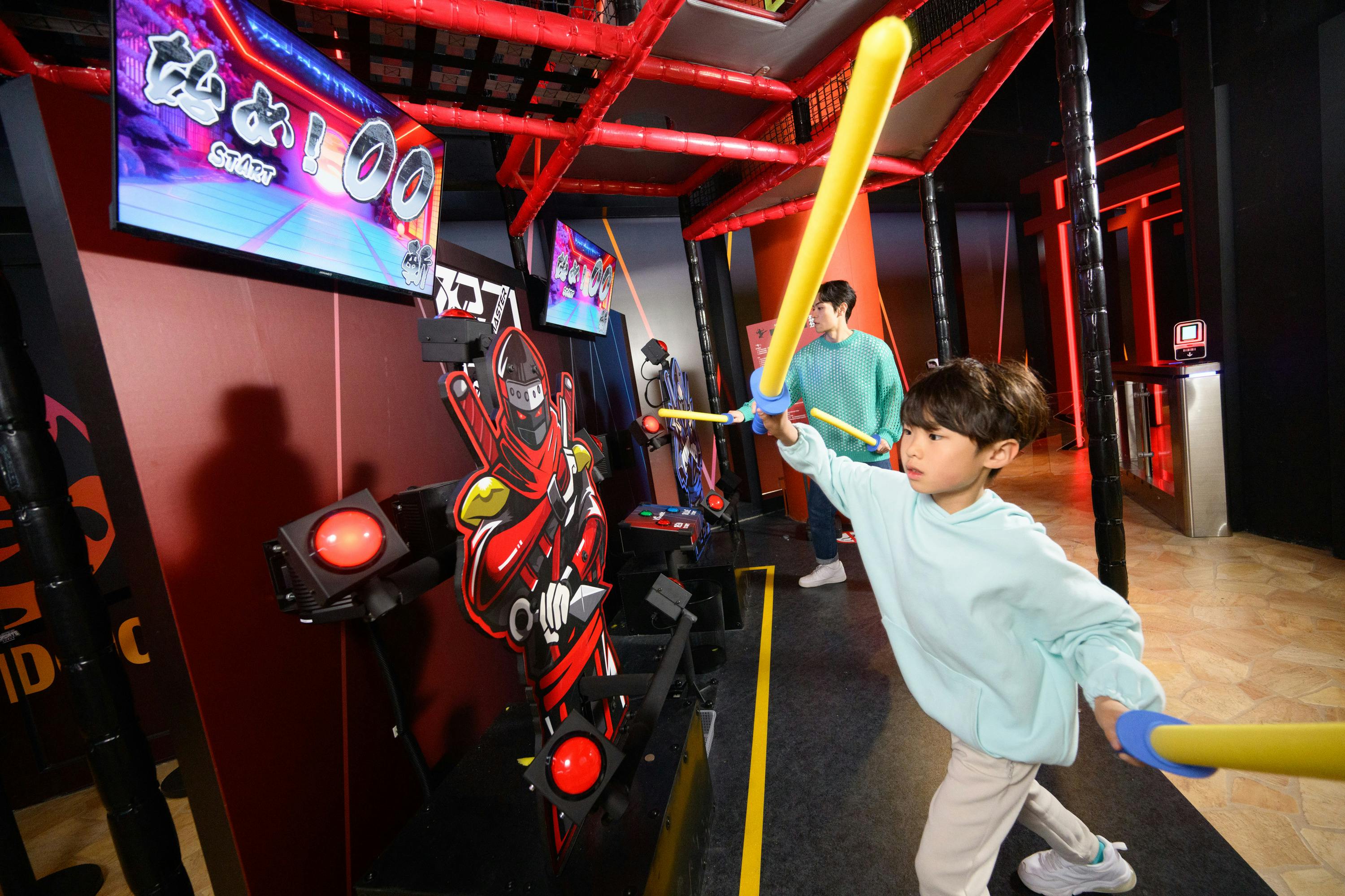 A child and an adult are playing a game with foam swords in an indoor activity center with red and black decor.