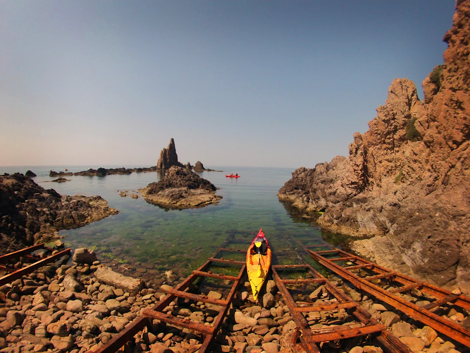 A person in a yellow kayak on a rocky shore, with calm seas and large rock formations in the background; another kayak can be seen in the distance.