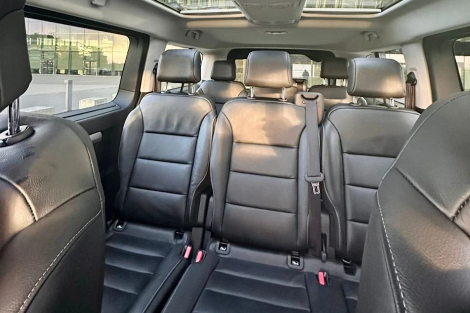 Interior of a car featuring three rows of black leather seats with visible seat belts and large windows.