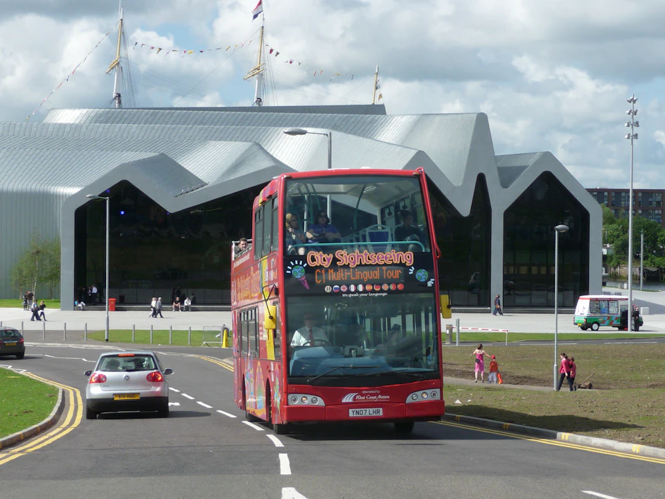 A red double-decker tour bus driving on a road near a modern building with a wavy roof, with cars and pedestrians nearby.