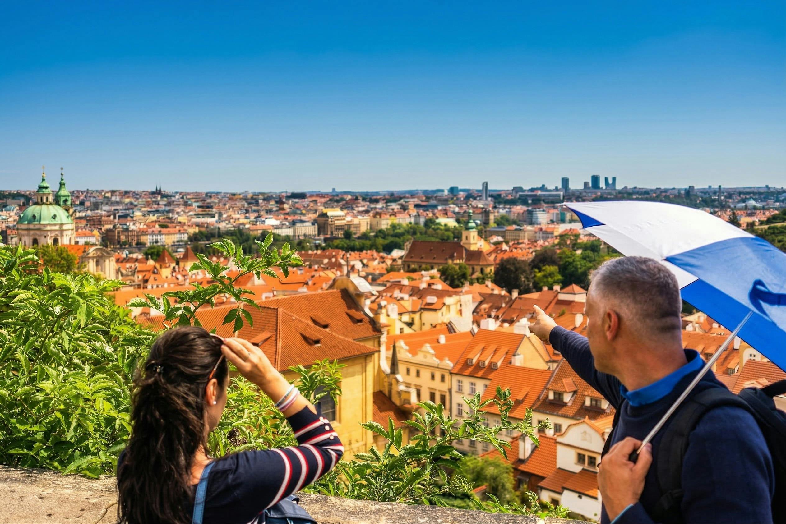 Twee mensen kijken uit over een schilderachtig stadsgezicht met rode daken onder een strakblauwe hemel, de een wijst naar het uitzicht.