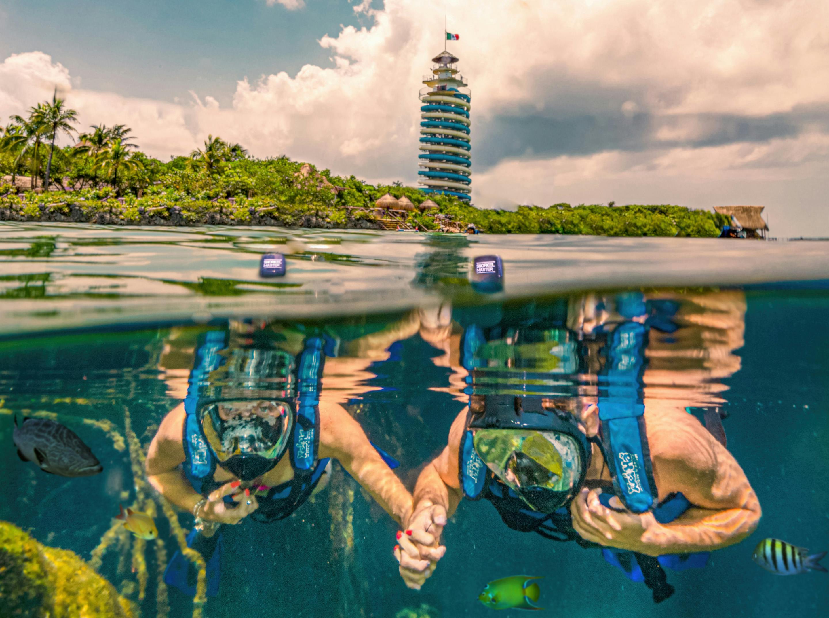 Deux plongeurs en apnée sous l'eau près d'une île verte avec une tour d'observation à plusieurs étages en arrière-plan sous un ciel nuageux.