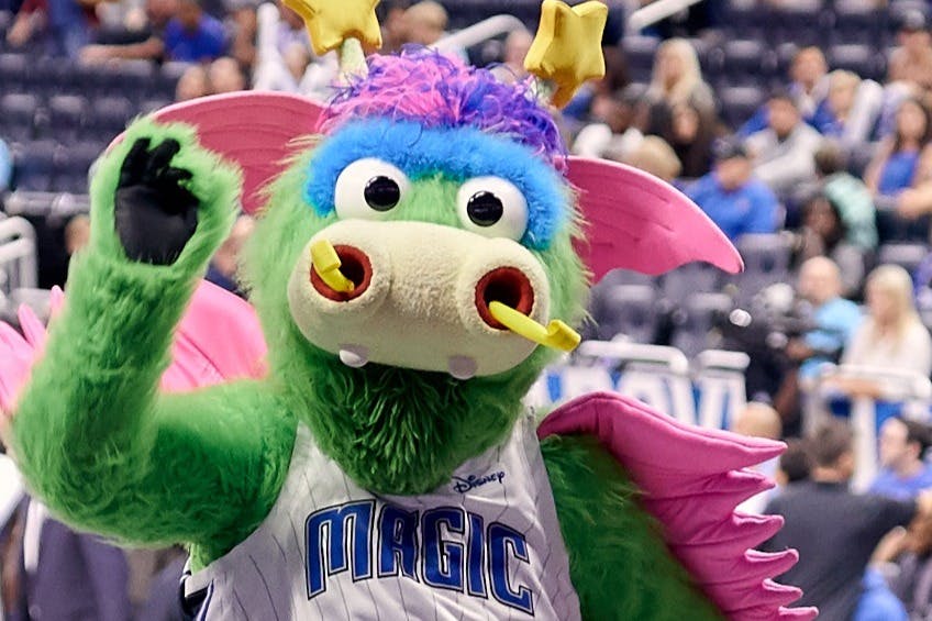 Μια πράσινη, πολύχρωμη μασκότ με μεγάλα μάτια και φτερά σε μια αθλητική εκδήλωση, με θολούς θεατές στο παρασκήνιο.