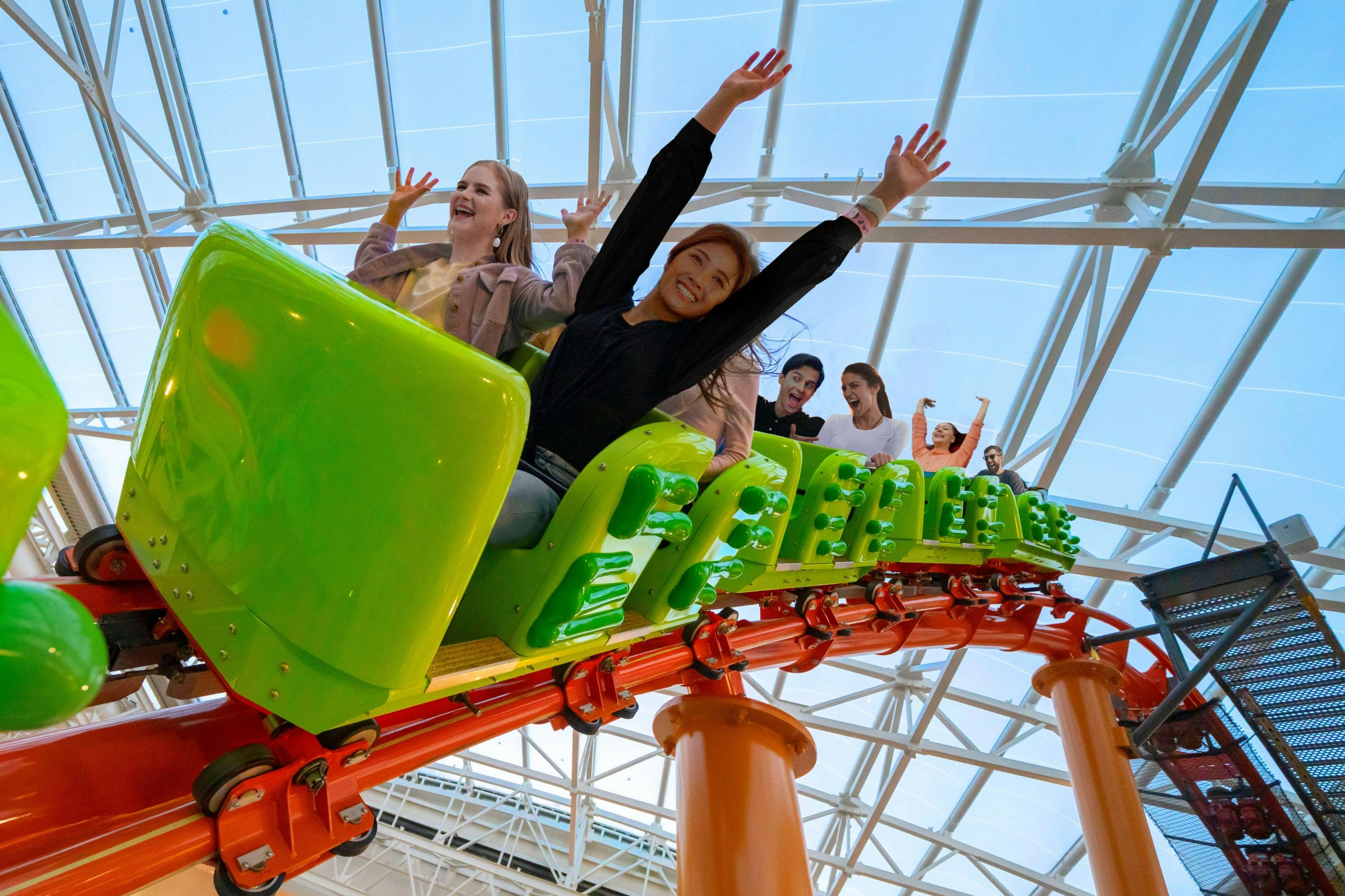 People riding a green rollercoaster indoors, smiling and raising their hands in excitement.