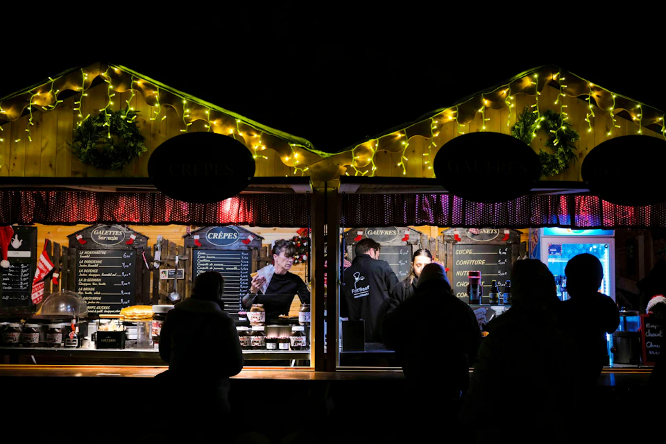 Two food stalls are lit up at night with people placing orders; one is labelled ‘Crêpes’ and the other sells a variety of toppings.